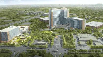 Aerial view of a modern hospital complex with multiple buildings surrounded by greenery and roads under a clear sky.