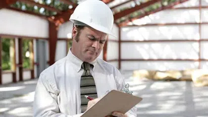Engineer wearing hard hat and tie inspecting construction site while writing on clipboard under steel frame.