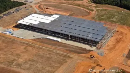 Aerial view of a large steel warehouse under construction surrounded by cleared land and earthmoving equipment.