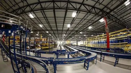 Wide view of an industrial warehouse with blue conveyor belts and yellow storage racks under a metal roof.
