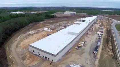 Aerial view of a large industrial warehouse under construction surrounded by dirt roads and forested areas.