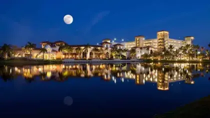 Luxury resort at night with bright lights reflecting on calm water and a full moon in the clear sky.