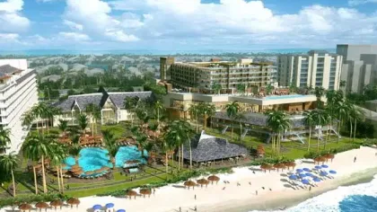Resort with multiple pools, palm trees, beach umbrellas, and beachfront buildings under a blue sky with clouds.