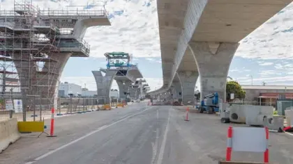Construction site showing elevated concrete bridge supports and scaffolding under a cloudy sky.