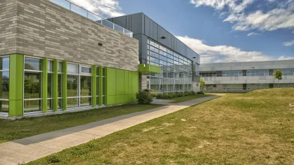 Modern building with green accents, glass windows, and a pathway beside a grassy area under a partly cloudy sky