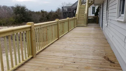 Wide wooden deck with railing attached to a house, stairs leading to an upper level, trees and grass in background.