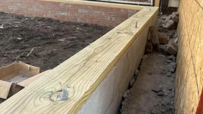 Concrete block wall with wooden cap and earth trench during outdoor construction near brick wall.