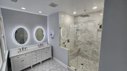 Modern bathroom with double sinks, illuminated oval mirrors, glass shower, and hexagonal floor tiles