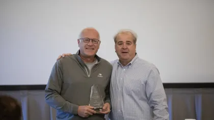 Two men smiling, one holding a clear glass award in a casual indoor setting with a coffee cup nearby.