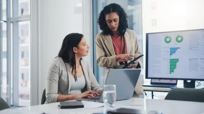 Two businesswomen discussing data charts on a tablet and laptop in a modern office with large windows.
