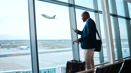 Businessman with suitcase checking phone while watching airplane take off at airport terminal window.