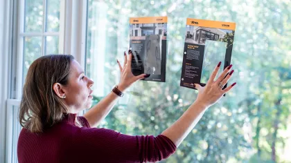 Woman comparing two transparent window film samples against a large window with trees outside.