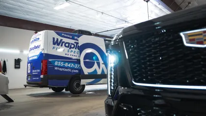 Blue and white Wraptastic van inside a garage with a black Cadillac vehicle close to the camera.