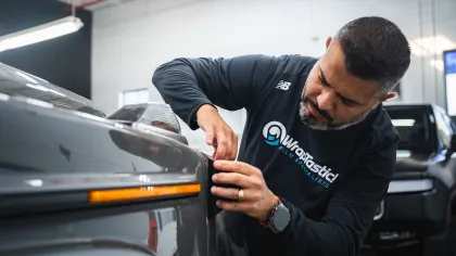 Technician applying window film to a vehicle's front headlight in a workshop with focused attention.