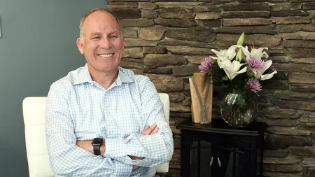 Smiling man in checkered shirt sitting with folded arms beside a vase of flowers against a stone wall indoors
