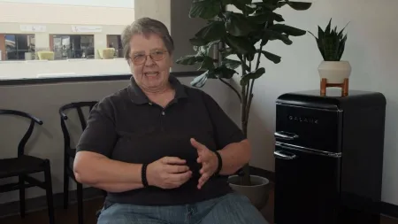 Person in black polo and jeans sitting indoors near a small black Galanz fridge and potted plants.