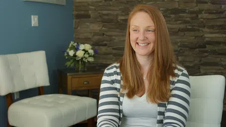 Smiling woman with long red hair in striped cardigan sitting in a cozy room with stone wall and white chairs.