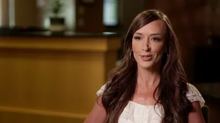 Smiling woman with long brown hair and hoop earrings sitting indoors in a warmly lit room.