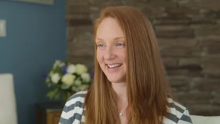 Smiling red-haired woman in striped shirt sitting indoors with blurred flowers and stone wall background