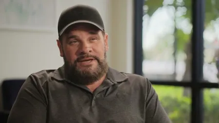 Man with a beard wearing a black cap and polo shirt, speaking indoors near a window with greenery outside.