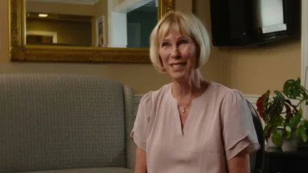 Smiling middle-aged woman sitting on a sofa in a warmly lit living room with plants and a large mirror.