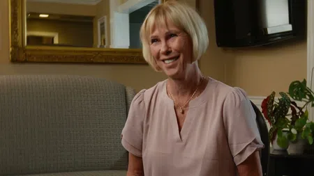Smiling middle-aged woman with short blonde hair sitting on a sofa in a warmly lit room with plants and a mirror.