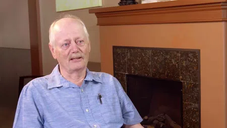 Elderly man in a light blue shirt speaking while seated near a fireplace with brown tiles.