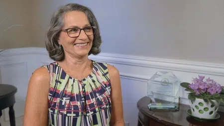 Smiling middle-aged woman with glasses and gray hair wearing a patterned sleeveless top indoors beside a table with award and flowers