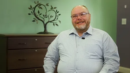 Smiling man with glasses and beard wearing checkered shirt sitting in office with tree decoration behind him.