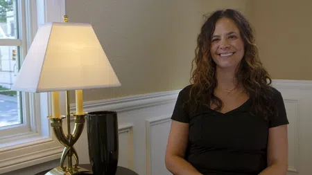 Smiling woman with curly hair sitting indoors near a table lamp and window in a bright room.