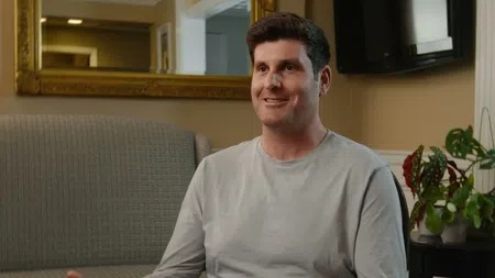 Man in light gray long sleeve shirt sitting and smiling in a warmly lit living room with a gold framed mirror and plants.