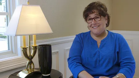 Smiling woman in blue sweater sitting next to a table lamp in a cozy, well-lit room with beige walls.