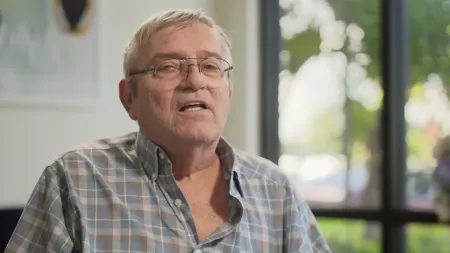 Older man wearing glasses and a plaid shirt speaking indoors with a window view of greenery behind him.