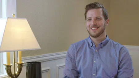 Smiling man with short hair and beard wearing a light blue button-up shirt sitting by a lamp in a neutral-toned room
