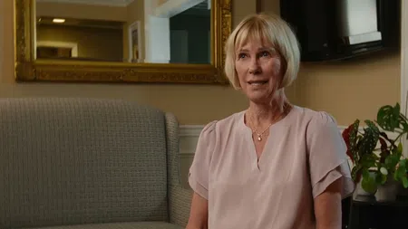 Blonde woman in a light pink blouse sits on a gray sofa in a warmly lit living room with plants and a large mirror.