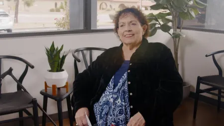 Older woman with curly hair wearing a patterned top and black jacket sitting in a bright room with plants and chairs.