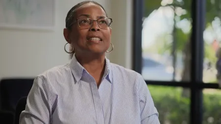 Smiling woman with glasses and hoop earrings wearing a striped shirt indoors near a window with natural light.
