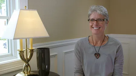 Smiling woman with short gray hair and glasses sitting by a window next to a gold lamp and black vase.