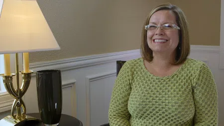 Smiling woman with glasses in green sweater sitting by a lamp and black vase in a cozy room.