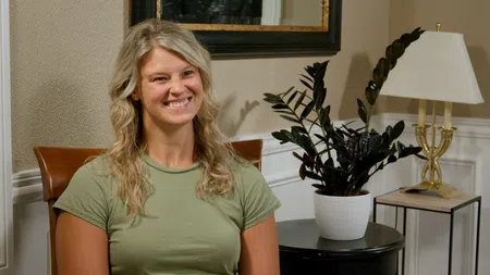 Smiling woman with blonde hair wearing green shirt sitting indoors with plant and lamp in background