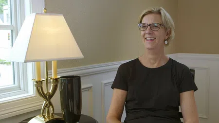 Smiling woman with glasses sitting indoors next to a lit table lamp and window during daytime