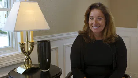 Smiling woman in black sitting near a table with a gold lamp and black vase by a window in a bright room.
