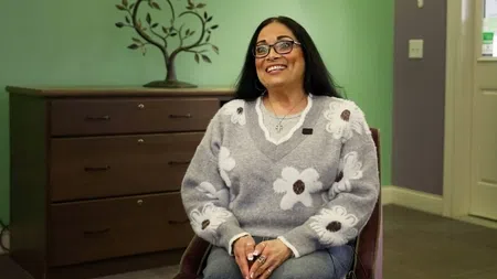 Smiling woman with glasses wearing a gray flower-patterned sweater sitting in a chair in a cozy room.