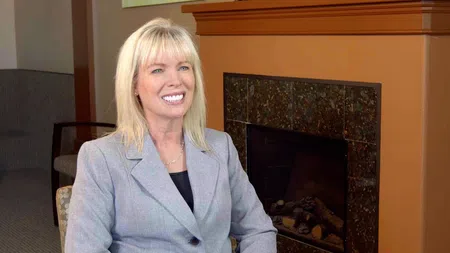 Smiling blonde woman in gray blazer seated near a modern fireplace in a warmly lit room.