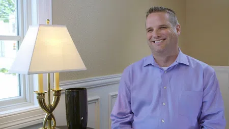 Smiling man in lavender shirt sitting next to a lit table lamp by a window in a cozy room.