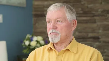 Elderly man with white hair and beard wearing a yellow shirt, sitting indoors with blurred flowers in background.