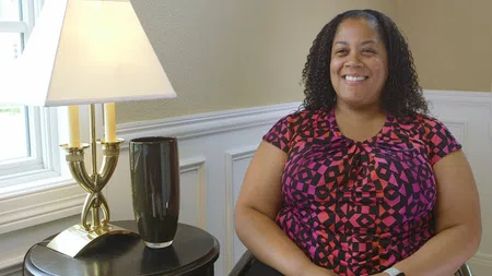 Smiling woman in patterned pink and black shirt sitting next to a round table with lamp and vase by window.