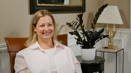 Smiling woman sitting on a chair in a cozy room with a plant and lamp in the background
