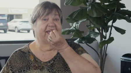 Elderly woman wearing patterned shirt eating a small snack indoors near a large leafy plant.