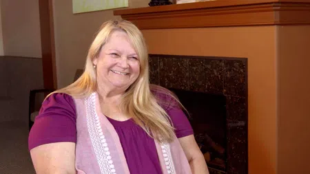 Smiling middle-aged woman with long blonde hair wearing a purple top and pink scarf sitting by a fireplace.